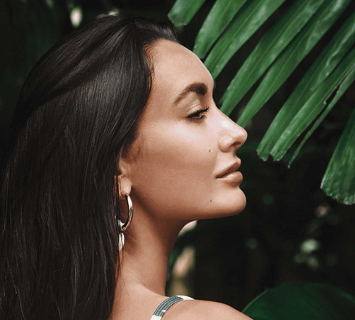 Woman in profile with long dark hair beside green palm leaves